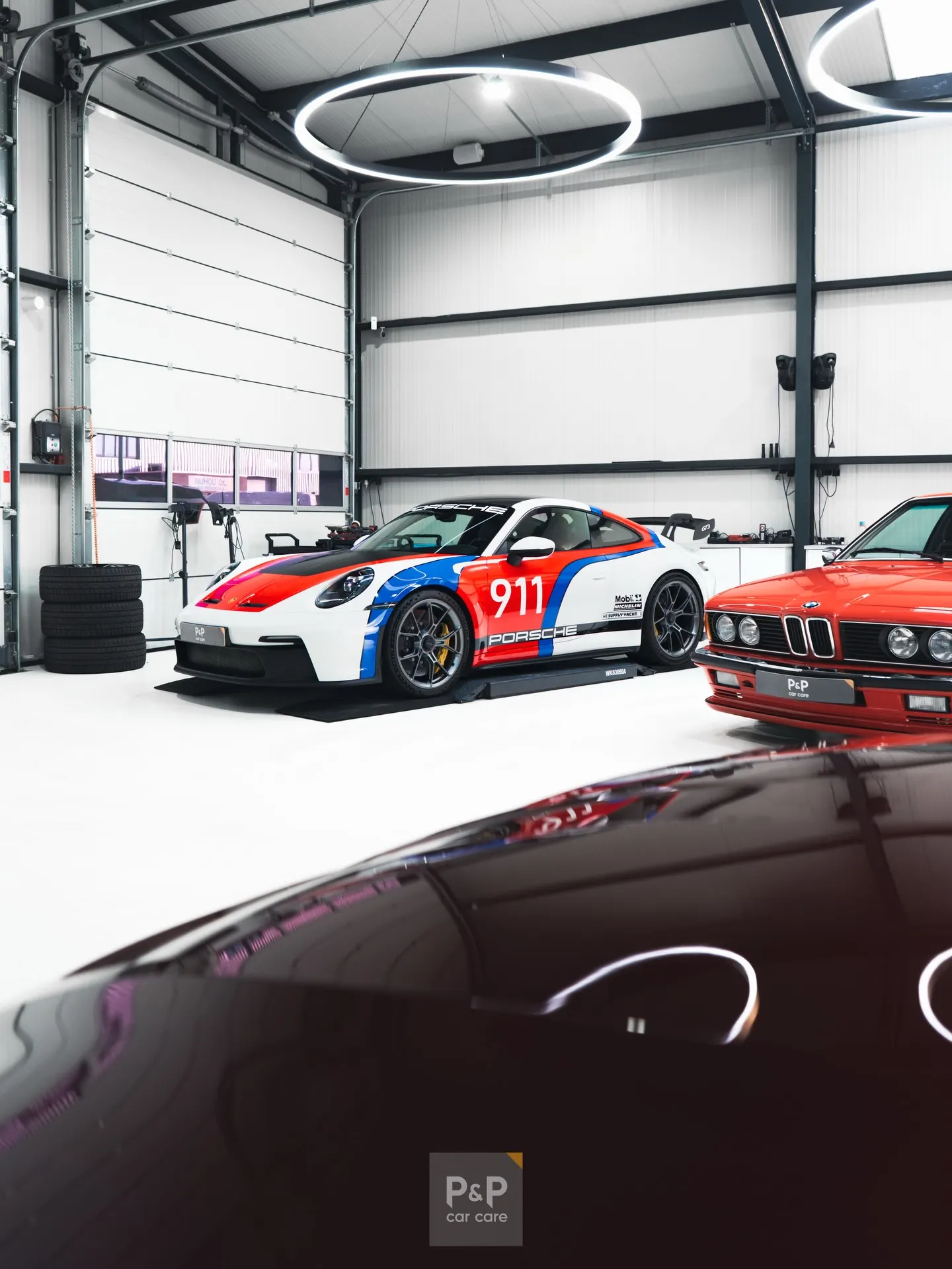 Witte Porsche 911 GT3 in een rood-blauwe racelivery in de werkplaats van P&P Car Care, naast een rode BMW 635 CSi
